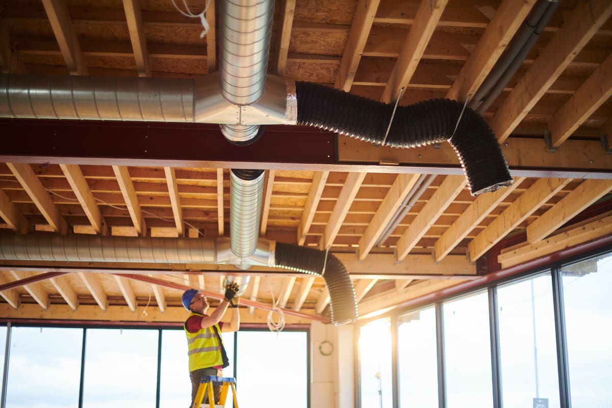 Worker installing ductwork on an exposed wooden ceiling.